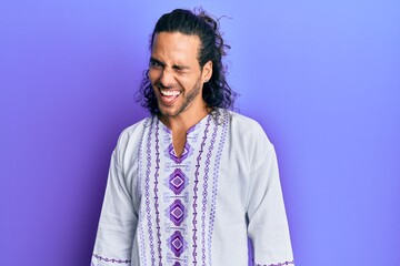 Young handsome man with long hair wearing bohemian and hippie shirt winking looking at the camera...