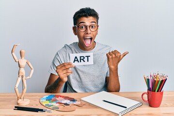 Naklejka premium Young handsome african american man painter sitting at art studio holding art word pointing thumb up to the side smiling happy with open mouth