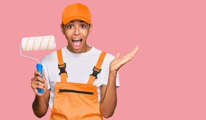Young handsome african american man wearing cap and painter clothes holding painting roll celebrating victory with happy smile and winner expression with raised hands