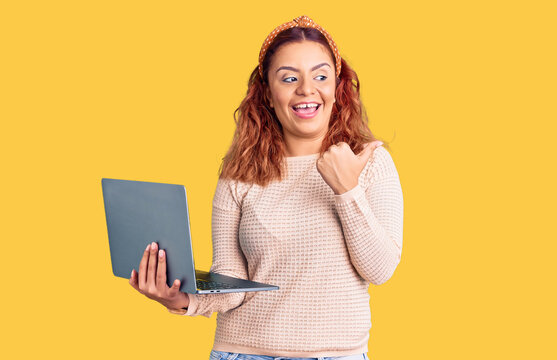 Young latin woman holding laptop pointing thumb up to the side smiling happy with open mouth