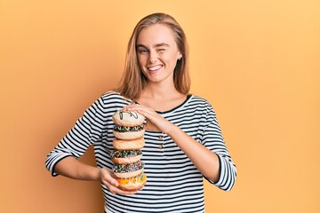 Beautiful blonde woman holding tasty colorful doughnuts winking looking at the camera with sexy...