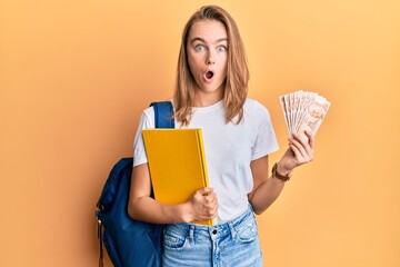 Beautiful blonde woman wearing student backpack and holding 50 turkish lira in shock face, looking...