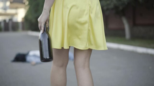 Camera follows unrecognizable drunk Caucasian woman with wine bottle walking to corpse hit by vehicle. Slim young female driver causing fatal road accident. Drunken driving concept.