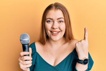 Young beautiful redhead woman singing song using microphone smiling with an idea or question pointing finger with happy face, number one