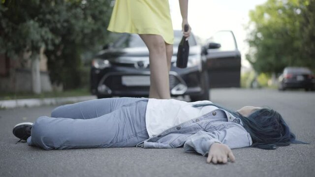 Road accident victim lying on asphalt as drunk young woman approaching to body holding wine bottle. Unrecognizable drunken Caucasian driver and corpse hit by car. Alcohol abuse and driving.