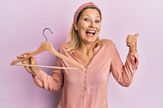 Middle age caucasian woman holding clothes hanger pointing thumb up to the side smiling happy with open mouth