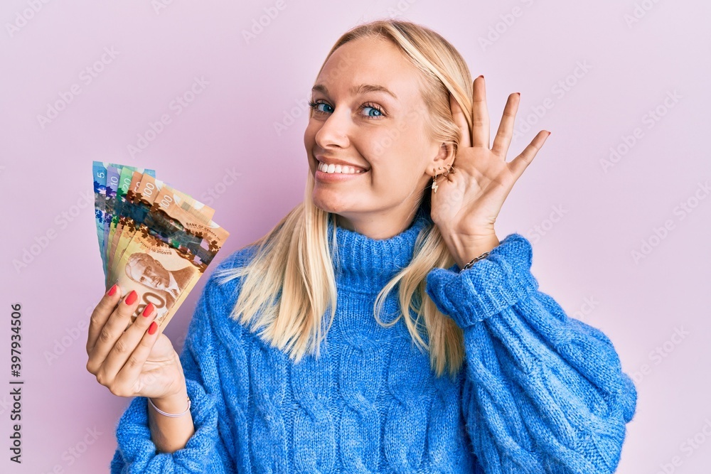 Wall mural Young blonde girl holding canadian dollars smiling with hand over ear listening an hearing to rumor or gossip. deafness concept.