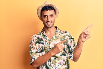 Young hispanic man wearing summer hat smiling and looking at the camera pointing with two hands and fingers to the side.