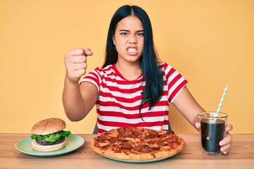 Young beautiful asian girl eating fast food pizza and burger annoyed and frustrated shouting with anger, yelling crazy with anger and hand raised