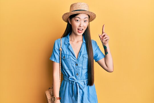Young chinese woman wearing summer hat pointing finger up with successful idea. exited and happy. number one.