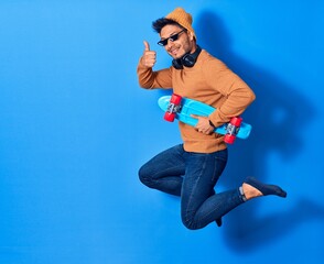 Young handsome latin man wearing wool cap and thug life glasses.  Jumping with smile on face holding skate over isolated blue background