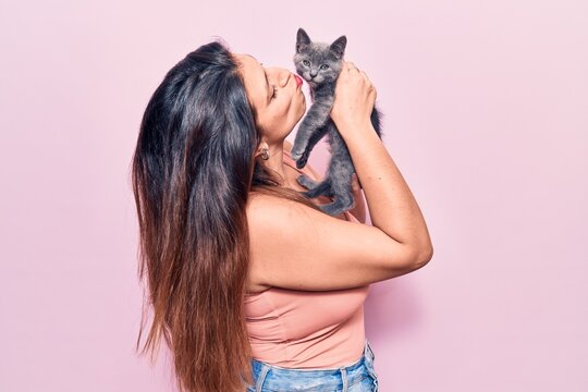 Young beautiful latin woman smilling happy. Standing with smile on face holding and kissing adorable cat over isolated pink background