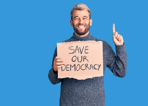 Young blond man holding save our democracy cardboard banner surprised with an idea or question pointing finger with happy face, number one