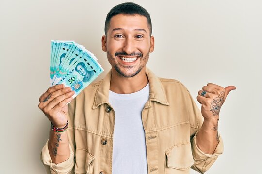 Handsome man with tattoos holding 50 yuan chinese banknotes pointing thumb up to the side smiling happy with open mouth