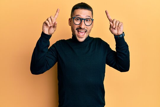 Handsome man with tattoos wearing turtleneck sweater and glasses smiling amazed and surprised and pointing up with fingers and raised arms.