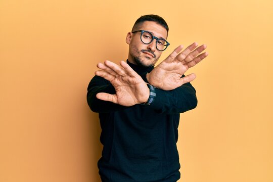 Handsome man with tattoos wearing turtleneck sweater and glasses rejection expression crossing arms and palms doing negative sign, angry face