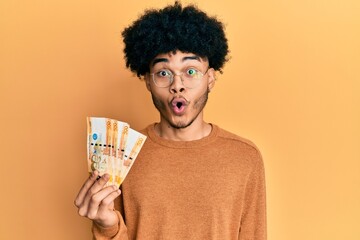 Young african american man with afro hair holding 500 philippine peso banknotes scared and amazed with open mouth for surprise, disbelief face