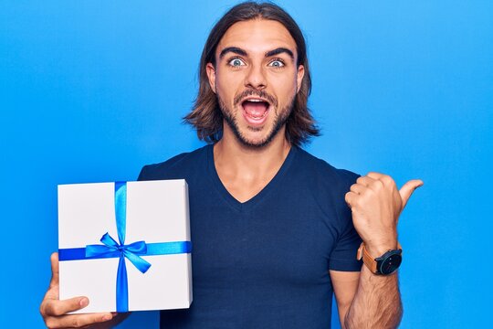 Young handsome man holding gift pointing thumb up to the side smiling happy with open mouth