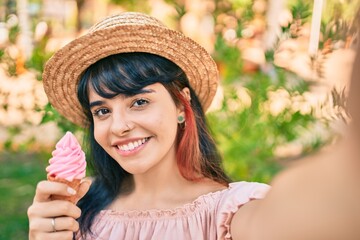 Young hispanic tourist girl wearing summer style eating ice cream and making selfie by the camera at the park.