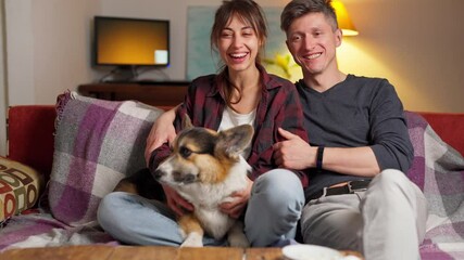 happy couple with cute pet Welsh Corgi dog sitting on couch at home, enjoying by spending time together and watching TV at evening smiling. Togetherness and entertainment.