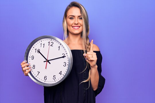 Young beautiful blonde woman doing countdown using big clock over purple background smiling with an idea or question pointing finger with happy face, number one