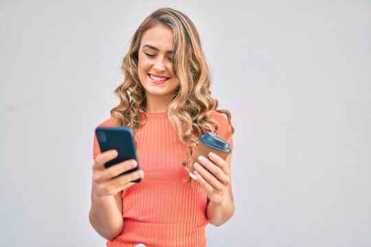 Young blonde girl smiling happy using smartphone and drinking take away coffee at the city.