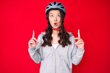Young beautiful chinese girl wearing bike helmet amazed and surprised looking up and pointing with fingers and raised arms.
