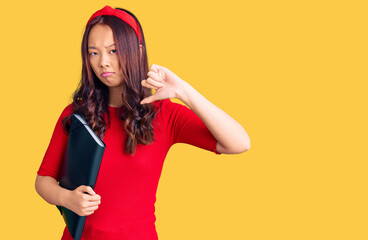 Young beautiful chinese girl wearing student folder with angry face, negative sign showing dislike with thumbs down, rejection concept