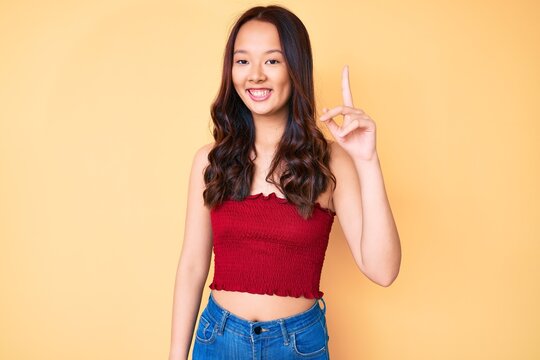 Young beautiful chinese girl wearing summer clothes pointing finger up with successful idea. exited and happy. number one.