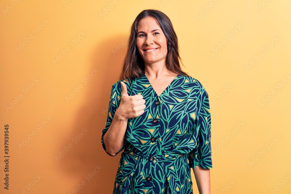 Wall mural young beautiful brunette woman wearing casual floral dress standing over yellow background doing hap