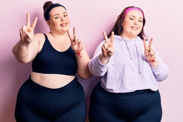 Young plus size twins wearing sportswear smiling looking to the camera showing fingers doing...
