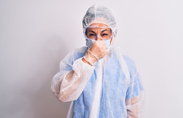 Middle age nurse woman wearing protection coronavirus equipment over white background smelling something stinky and disgusting, intolerable smell, holding breath with fingers on nose. Bad smell