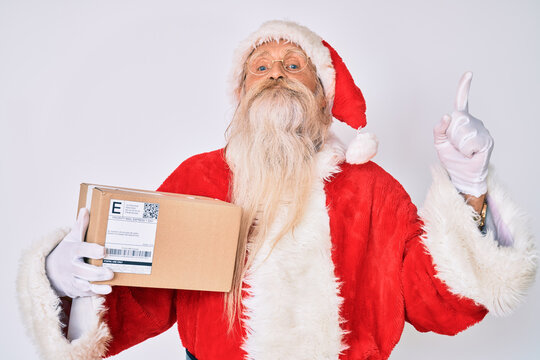 Old senior man with grey hair and long beard wearing santa claus costume holding delivery box smiling with an idea or question pointing finger with happy face, number one
