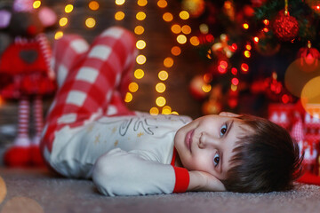 Obraz na płótnie Canvas A beautiful, cute boy in Christmas pajamas is lying on the background of a Christmas tree and garland lights. Gifts under the tree.