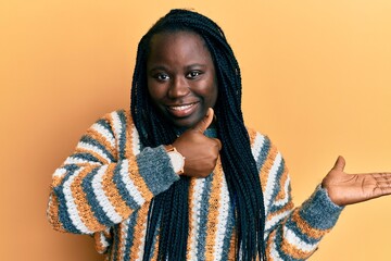 Young black woman with braids wearing casual winter sweater showing palm hand and doing ok gesture with thumbs up, smiling happy and cheerful