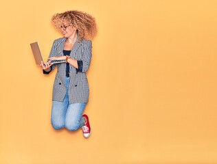 Young beautiful curly businesswoman wearing glasses smiling happy. Jumping with smile on face working using laptop over isolated yellow background.