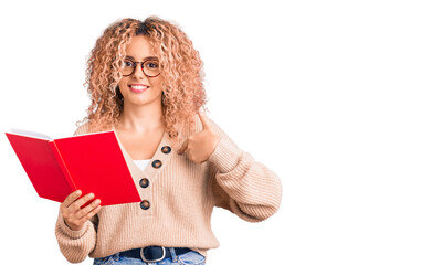 Young blonde woman with curly hair wearing glasses and reading book pointing finger to one self smiling happy and proud