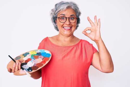 Senior hispanic grey- haired woman holding paintbrush and palette doing ok sign with fingers, smiling friendly gesturing excellent symbol