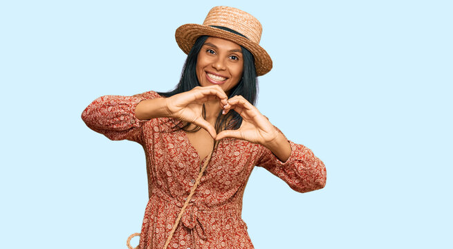 Young african american woman wearing summer hat smiling in love doing heart symbol shape with hands. romantic concept.