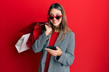 Young brunette woman holding shopping bags and smartphone smiling looking to the side and staring away thinking.