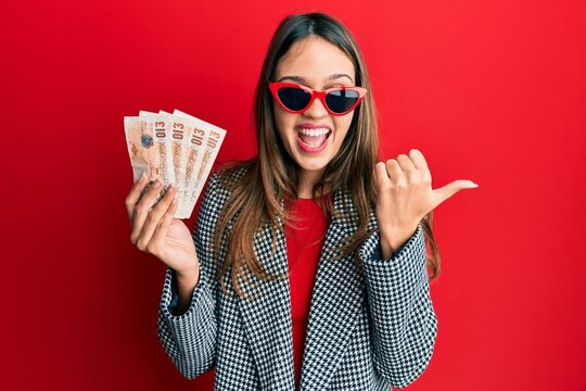 Young brunette woman holding united kingdom 10 pounds banknotes pointing thumb up to the side smiling happy with open mouth
