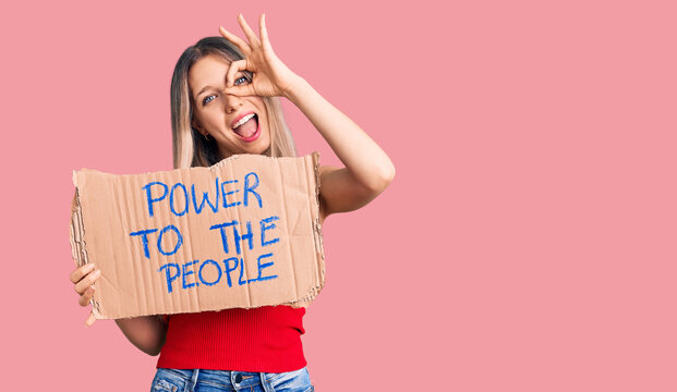Young beautiful blonde woman holding power to the people banner smiling happy doing ok sign with hand on eye looking through fingers