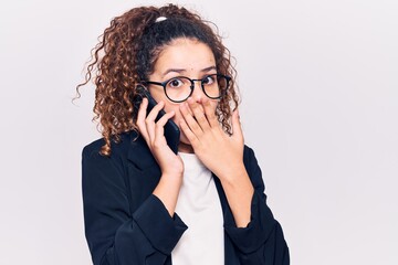 Beautiful kid girl with curly hair wearing glasses having conversation talking on the smartphone covering mouth with hand, shocked and afraid for mistake. surprised expression