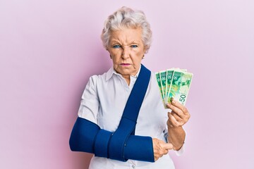 Senior grey-haired woman wearing arm on sling holding israel shekels banknotes depressed and worry...