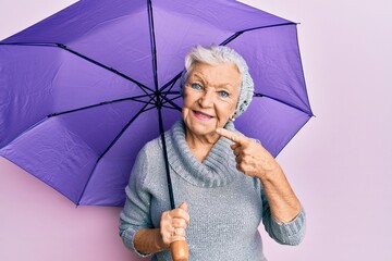 Senior grey-haired woman holding purple umbrella smiling happy pointing with hand and finger