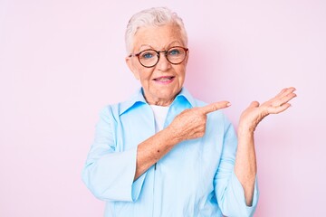 Senior beautiful woman with blue eyes and grey hair wearing glasses amazed and smiling to the camera while presenting with hand and pointing with finger.