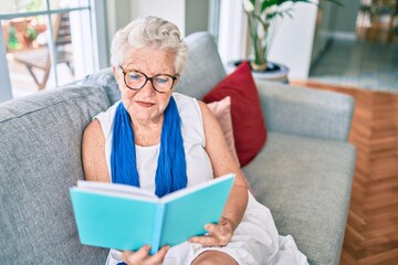 Elder senior woman with grey hair smiling happy sitting on the sofa reading a book at home