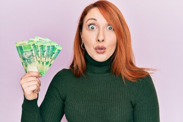 Beautiful redhead woman holding south african 10 rand banknotes scared and amazed with open mouth for surprise, disbelief face