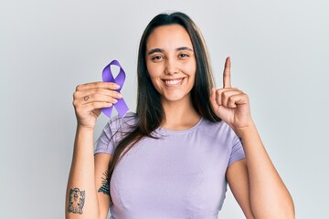 Young hispanic woman holding purple ribbon awareness surprised with an idea or question pointing...