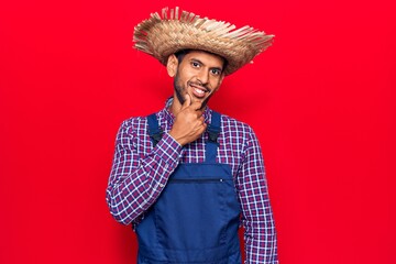 Young latin man wearing farmer hat and apron looking confident at the camera with smile with crossed arms and hand raised on chin. thinking positive.